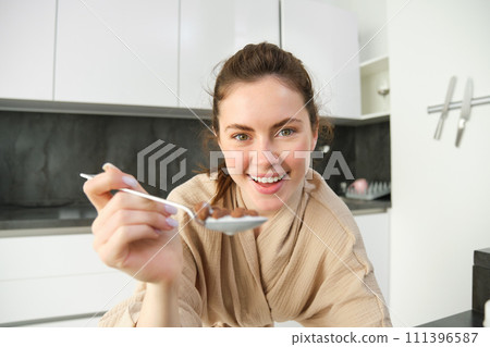 Portrait of happy young woman leans on kitchen worktop and eating cereals, has milk and bowl in front of her, having her breakfast, wearing bathrobe Portrait of happy young woman leans on kitchen worktop and eating cereals, has milk and bowl in front of her, having her breakfast, wearing bathrobe 111396587