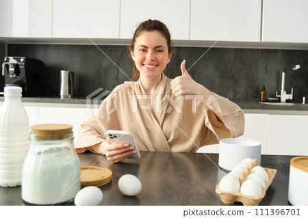 Attractive young cheerful girl baking at the kitchen, making dough, holding recipe book, having ideas 111396701