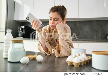 Attractive young cheerful girl baking at the kitchen, making dough, holding recipe book, having ideas Attractive young cheerful girl baking at the kitchen, making dough, holding recipe book, having ideas 111396719