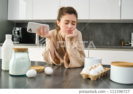 Attractive young cheerful girl baking at the kitchen, making dough, holding recipe book, having ideas 111396727