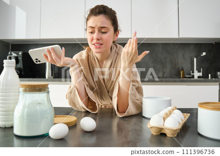 Attractive young cheerful girl baking at the kitchen, making dough, holding recipe book, having ideas Attractive young cheerful girl baking at the kitchen, making dough, holding recipe book, having ideas 111396736