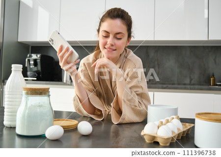 Attractive young cheerful girl baking at the kitchen, making dough, holding recipe book, having ideas 111396738