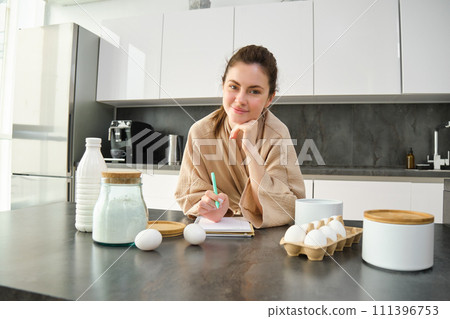 Attractive young cheerful girl baking at the kitchen, making dough, holding recipe book, having ideas 111396753
