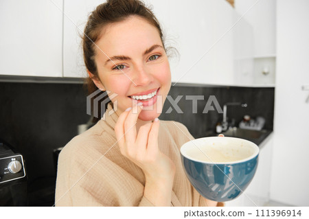 Close up portrait of attractive girl drinking coffee, holding cup with morning cappuccino and smiling, having a mug of delicious drink in the kitchen, wearing comfortable bathrobe Close up portrait of attractive girl drinking coffee, holding cup with morning cappuccino and smiling, having a mug of delicious drink in the kitchen, wearing comfortable bathrobe 111396914