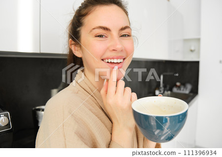 Close up portrait of attractive girl drinking coffee, holding cup with morning cappuccino and smiling, having a mug of delicious drink in the kitchen, wearing comfortable bathrobe Close up portrait of attractive girl drinking coffee, holding cup with morning cappuccino and smiling, having a mug of delicious drink in the kitchen, wearing comfortable bathrobe 111396934