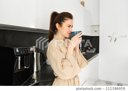 Portrait of good-looking young woman starting her day with cup of coffee, standing in the kitchen and drinking cappuccino from big mug, enjoying favourite drink in the morning 111396959