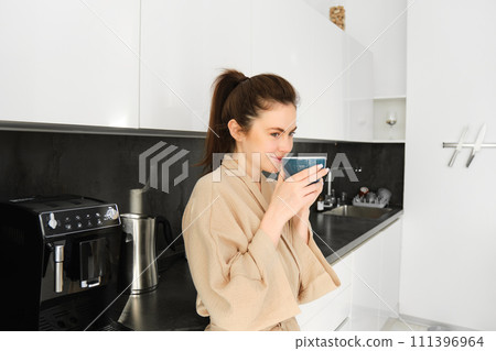 Portrait of woman standing near coffee machine, preparing morning cup of cappuccino, standing in kitchen, wearing bathrobe 111396964