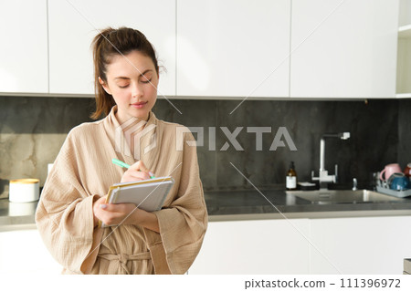 Portrait of woman writing grocery list for shopping, standing in the kitchen and making notes for recipe, cooking at home in bathrobe Portrait of woman writing grocery list for shopping, standing in the kitchen and making notes for recipe, cooking at home in bathrobe 111396972