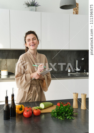 Vertical shot of young woman writing down food ideas, creating new meal, writing down grocery list or recipe in the notebook, standing in the kitchen near chopping board with fresh vegetables 111396975