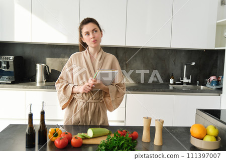 Portrait of creative young woman, cooking, holding recipe book, thinking while making meal, preparing food in the kitchen, standing in bathrobe Portrait of creative young woman, cooking, holding recipe book, thinking while making meal, preparing food in the kitchen, standing in bathrobe 111397007