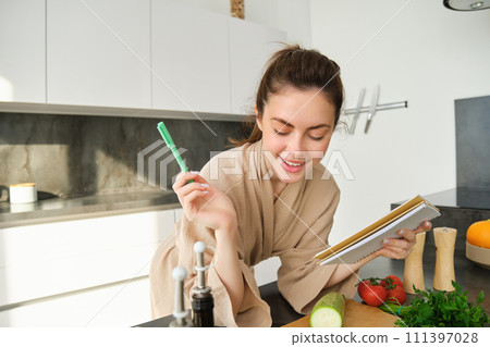 Portrait of woman checking grocery list, looking at vegetables, holding notebook, reading recipe while cooking meal in the kitchen, chopping tomatoes and zucchini 111397028