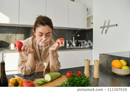 Portrait of woman cooking at home in the kitchen, holding tomatoes, preparing delicious fresh meal with vegetables, standing near chopping board Portrait of woman cooking at home in the kitchen, holding tomatoes, preparing delicious fresh meal with vegetables, standing near chopping board 111397038