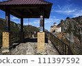 A gazebo with a bell on a rock, against the backdrop of the Shaonin Temple. View from the south. Village named after Kosta Khetagurov, Karachay district. Karachay-Cherkessia. Russia 111397592