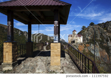 A gazebo with a bell on a rock, against the backdrop of the Shaonin Temple. View from the south. Village named after Kosta Khetagurov, Karachay district. Karachay-Cherkessia. Russia 111397592