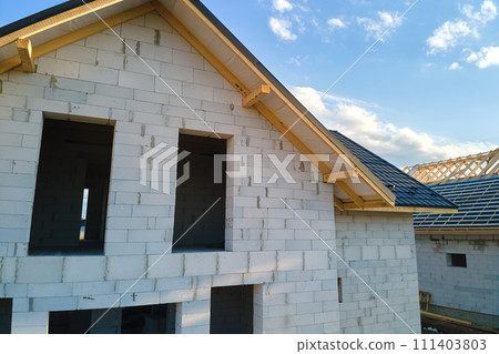 Aerial view of unfinished house with aerated lightweight concrete walls and wooden roof frame covered with metallic tiles under construction. Aerial view of unfinished house with aerated lightweight concrete walls and wooden roof frame covered with metallic tiles under construction. 111403803