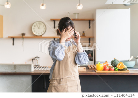 A housewife Asian woman crying with a headache, poor physical condition, and illness while holding a smartphone in the kitchen at home A housewife Asian woman crying with a headache, poor physical condition, and illness while holding a smartphone in the kitchen at home 111404134