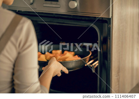 A young woman in an apron puts a dish with pumpkin in an electric oven for baking in the kitchen 111406407