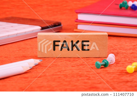 FAKE word written on a wooden block on an orange background next to office supplies 111407710