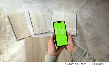 Hands Holding Device with Green Chroma Key Screen above Luxury Vinyl Wood Samples, Laminate On Floor In House. Choosing, Selecting Waterproof Flooring. Home Reconstruction. Horizontal Plane 111408903