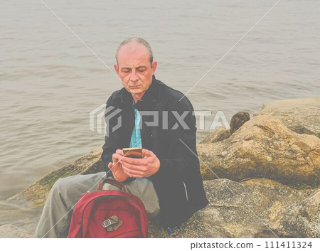 Midle aged man sitting on the shore of the lake. Solitude mature man using mobile phone on the bank. Concept of activ man Midle aged man sitting on the shore of the lake. Solitude mature man using mobile phone on the bank. Concept of activ man 111411324