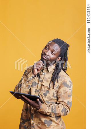 African american man browsing internet on digital tablet in studio over yellow background. Pensive young adult using wireless portable gadget surfing on social media app, watching video 111413284