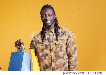 African american stylish man carrying shopping bags in hands after buying trendy clothes. Shopaholic young adult smiling at camera after purchase fashionable items in studio over yellow background African american stylish man carrying shopping bags in hands after buying trendy clothes. Shopaholic young adult smiling at camera after purchase fashionable items in studio over yellow background 111413289