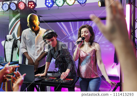 Dj and singer performing on stage at discotheque party in nightclub illuminated with colorful spotlights. Electronic music band singing and playing at disco gathering event in club Dj and singer performing on stage at discotheque party in nightclub illuminated with colorful spotlights. Electronic music band singing and playing at disco gathering event in club 111413293