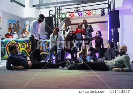 Music band performing on stage while diverse tired people lying and sitting on dancefloor. Exhausted young clubbers relaxing and listening to live music at club party gathering Music band performing on stage while diverse tired people lying and sitting on dancefloor. Exhausted young clubbers relaxing and listening to live music at club party gathering 111413297