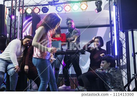 Electronic music band playing while clubbers relaxing and drinking near stage at club party. Dj performing at discotheque while young people resting on dancefloor in nightclub Electronic music band playing while clubbers relaxing and drinking near stage at club party. Dj performing at discotheque while young people resting on dancefloor in nightclub 111413300