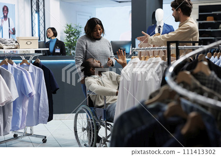 Man in wheelchair exploring clothing store and receiving guidance from mall assistant in selecting casual outfit. Shopping center customer with disability browsing through apparel rack Man in wheelchair exploring clothing store and receiving guidance from mall assistant in selecting casual outfit. Shopping center customer with disability browsing through apparel rack 111413302