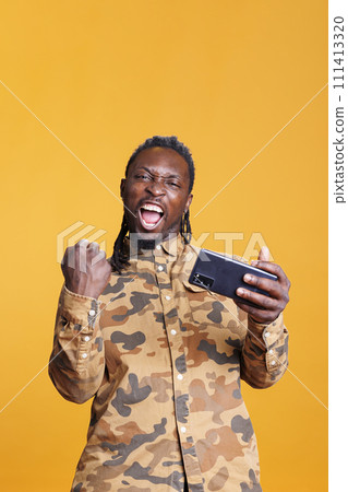 Joyful player winning online gaming tournament, enjoying playing video games during activity time. Competitive african american man having videogames competition in studio over yellow background 111413320