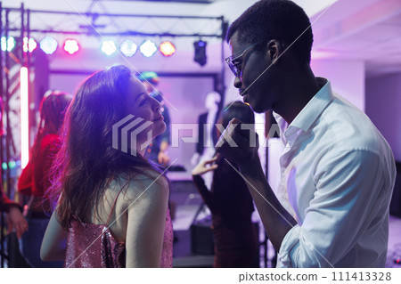 Couple in love dancing and clubbing while attending disco party in nightclub. Young diverse boyfriend and girfriend celebrating, moving to music rhythm on dancefloor illuminated with stage spotlights 111413328