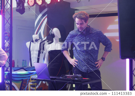 Dj standing on stage while mixing tracks with digital console at festival in nightclub. Young man using electronic music controller and performing at disco party event in club 111413383
