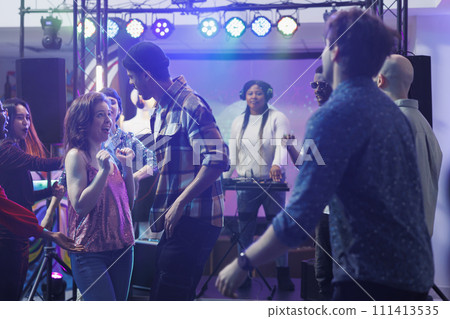 Young people dancing and partying at social gathering in nightclub with spotlights. Energetic clubbers relaxing on crowded dancefloor while clubbing at live music concert festival Young people dancing and partying at social gathering in nightclub with spotlights. Energetic clubbers relaxing on crowded dancefloor while clubbing at live music concert festival 111413535