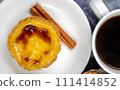 One Pastel de nata or Portuguese egg tart and cinnamon sticks on a white plate with a cup of coffee. Pastel de Belm is a small pie with a crispy puff pastry crust and a custard cream filling 111414852
