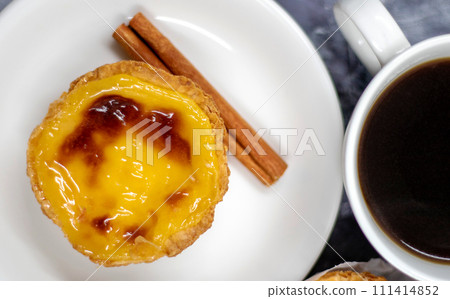 One Pastel de nata or Portuguese egg tart and cinnamon sticks on a white plate with a cup of coffee. Pastel de Belm is a small pie with a crispy puff pastry crust and a custard cream filling 111414852
