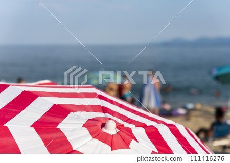 Striped beach umbrella at the beach with tourists and sea in background Striped beach umbrella at the beach with tourists and sea in background 111416276