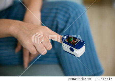 Young woman finger inserted into the pressure oximeter to assess health under sofa in living room at home, Self healthcare with corona virus pandemic, medical health care concept 111416516