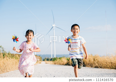 Brother and sister run by windmills holding pinwheels. Kids' joy near turbines represents a future of clean energy. Smiling children carefree and happy engage with sustainable technology. 111416570
