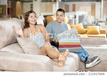 In store wife sit with legs on gray sofa to test in use,husband examines upholstery fabric In store wife sit with legs on gray sofa to test in use,husband examines upholstery fabric 111417849