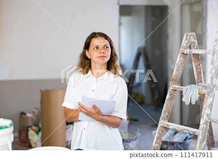 Portrait of positive woman next to a stepladder with documents in her hands checks the quality of the completed repairs in cottage room Portrait of positive woman next to a stepladder with documents in her hands checks the quality of the completed repairs in cottage room 111417911