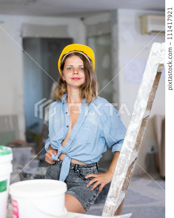 Seductive woman in hardhat and denim shirt posing next to a stepladder in room being renovated 111417921