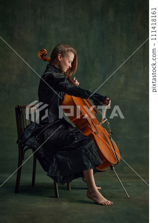 Passionate musician, elegant young woman, cellist in black dress sitting on chair and playing cello against dark vintage background. 111418161