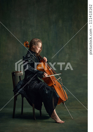 Tender young woman in black dress, cellist sitting on chair and playing cello against dark green background. Combination of elegance and music 111418162