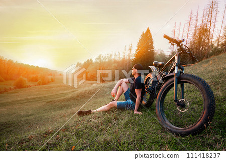 Man with an electric fatbike on the background of the sunset. Picturesque place in the village. Concept of a healthy lifestyle. 111418237