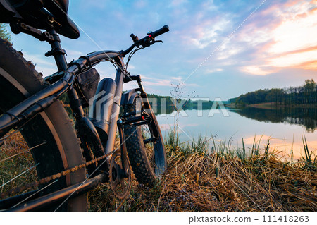Electric bike with thick wheels on the background of lake and sunset. Sports fatbike. Picturesque place in the village. The concept of a healthy lifestyle. 111418263