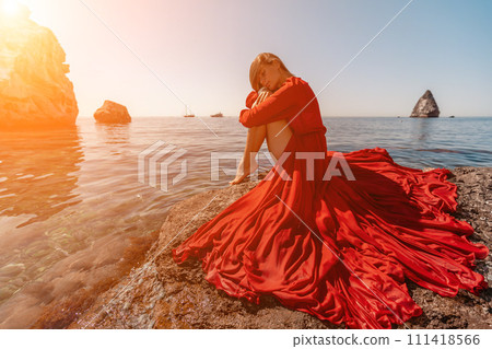 woman sea red dress. Beautiful sensual woman in a flying red dress and long hair, sitting on a rock above the beautiful sea in a large bay. woman sea red dress. Beautiful sensual woman in a flying red dress and long hair, sitting on a rock above the beautiful sea in a large bay. 111418566