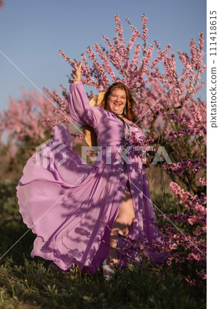 Woman blooming peach orchard. Against the backdrop of a picturesque peach orchard, a woman in a long pink dress and hat enjoys a peaceful walk in the park, surrounded by the beauty of nature. 111418615