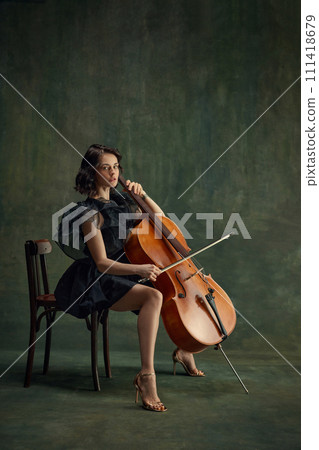 Pensive female musician, cellist sitting on chair with cello, in thoughtful pose against vintage green background. Poster for orchestra's seasonal performances Pensive female musician, cellist sitting on chair with cello, in thoughtful pose against vintage green background. Poster for orchestra's seasonal performances 111418679