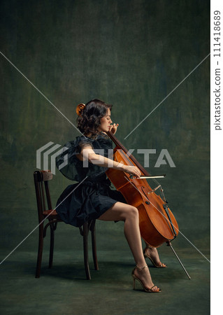 Attractive, elegant young woman, cellist, musician in black dress sitting on chair and playing cello against dark vintage background. 111418689